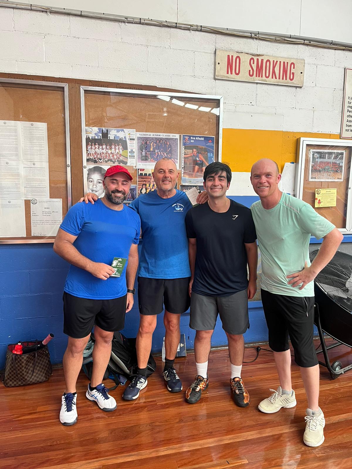 Four club members posing together by the courtside noticeboard after a session